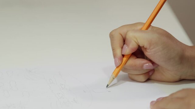 A close up shot of a woman's hand with manicured nails drawing a simple sketch on white paper using an orange pencil. The scene depicts a creative process, hobby, or art therapy session
