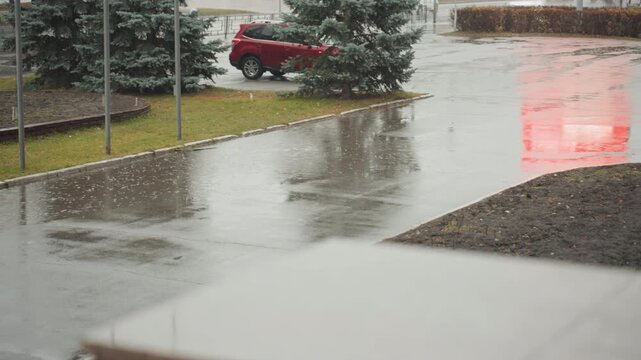 Wet entrance with red car nearby, slick driveway and puddles, falling sleet flakes, evergreen beside parking, overcast sky reflecting on asphalt, intimate observational tone for creator, commuter,