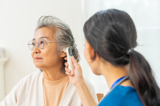 Asian female doctor examine senior woman patient with ear infrared thermometer measure temperature. Caregiver or nurse visit elderly patient health at home. Home medical healthcare service concept.