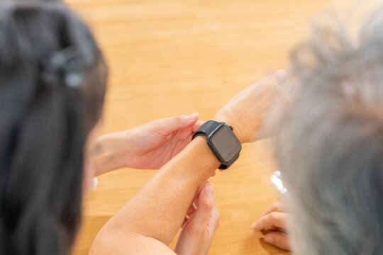 Adult daughter teaching senior mother use smartwatch at home, elderly learn digital technology, wearable device for health monitoring, family support, caregiving, and smart lifestyle for active aging.