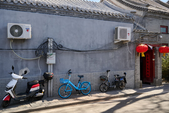 Electric scooters and bicycles parked along a gray brick wall with air conditioners and wiring in a Beijing Hutong, China, Asia.