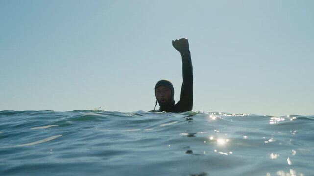 Surfer in wetsuit raising arm while floating in ocean water after wave ride, stoked with excitement and power