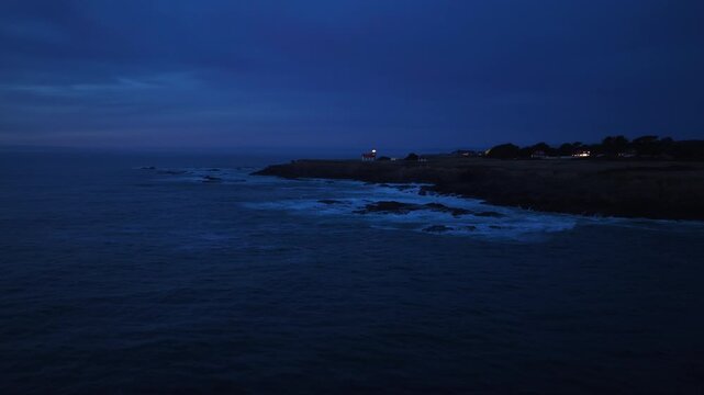 Night aerial view of Point Cabrillo Lighthouse in Mendocino, California, USA. Waves crash against rocky coastline as beacon light glows against deep blue twilight sky with distant buildings visible
