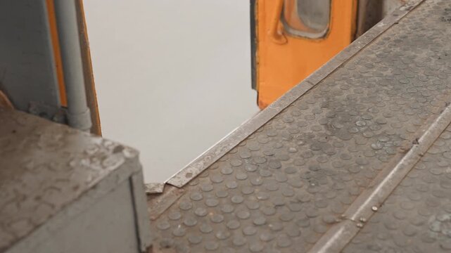 Orange train door platform gap closeup showing worn metal step and tactile floor plates beside orange subway door, peeling paint and rusted edges, gritty industrial texture, ambient urban atmosphere,