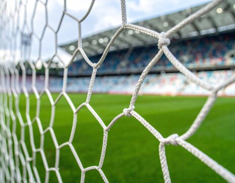 Detailed view through the white hexagonal mesh of a soccer goal net towards a blurred green football field and brightly colored stadium stands on a cloudy day, evoking sports and championship