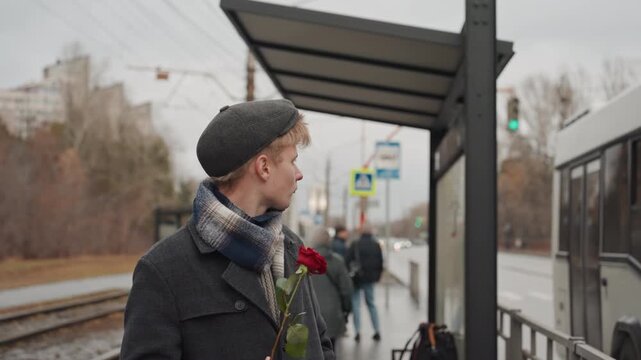 Young white creator at platform waiting anxious messenger vibe holding single red rose, coat and hat, distant stare down tracks, commuters passing, cool winter light, raw realistic atmosphere,