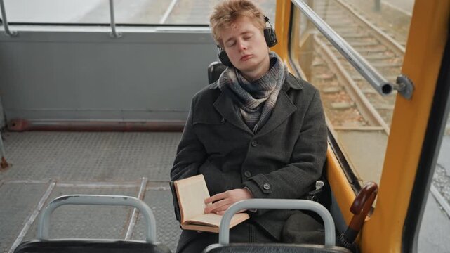 Young white creator writing in notebook. Headphones on, rails visible through window, focused expression, steady pen strokes on page, warm coat, creative planning, peaceful productive mood