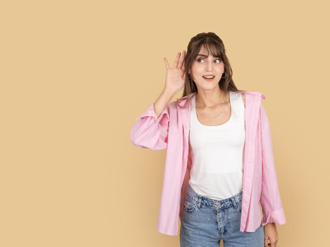 Curious caucasian woman holding hand near ear and listening carefully, reacting to interesting or surprising information, wearing pink shirt and denim, isolated on beige background with copy space.