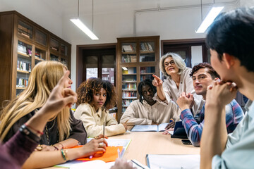 Group of diverse college students actively participating in a study session, discussing topics with a female professor in a library setting © Koldo_Studio