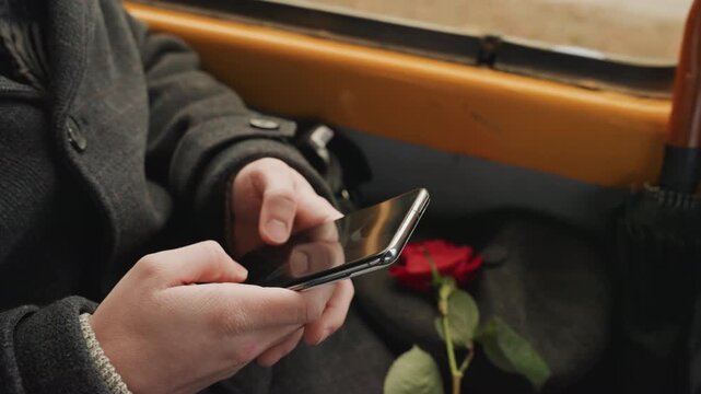 Hands holding smartphone with rose on lap, umbrella by seat, creator composing message, single red petal detail, wool coat sleeve, reflective window light, quiet romance mood, touchscreen scrolling,