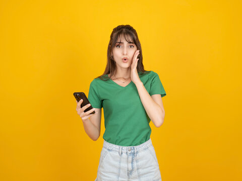 Surprised caucasian woman holding smartphone and touching face with shocked expression, reacting to unexpected message or news, wearing green t-shirt, isolated on yellow background with copy space.
