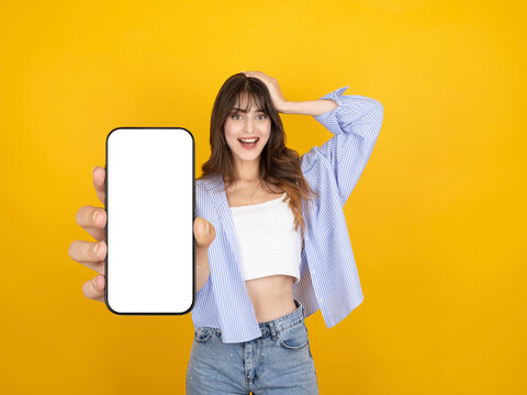 Excited caucasian woman showing smartphone with blank screen mock up toward camera, smiling with surprised expression, wearing striped shirt, isolated on yellow studio background with copy space.