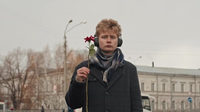 Young white creator holding red rose wearing headphones and wool coat, shifts roles from romantic giver to defiant protester to playful musician, facial closeups capture surprise, smirk and subtle