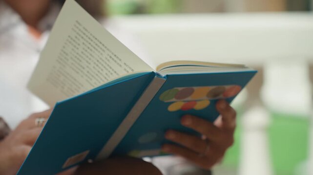 Lady enjoying book by coast. Sunlit veranda scene showing lady flipping through vibrant journal pages. Serene outdoor setting where woman relaxes on tropical porch with colorful magazine