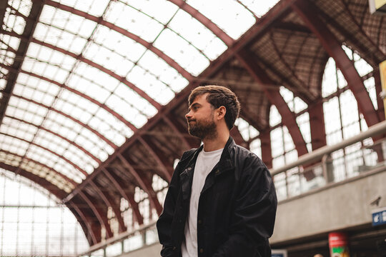 Disappointed young man traveler looking away with upset facial expression standing at train station after missing his departure or hearing bad news about trip on blurred architecture background