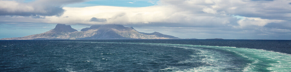 Fototapeta premium Island on the horizon. Rocks in the sea. Beautiful rocky sea landscape with dramatic cloudy sky. Water trail foaming behind a ferry. View from the ship. Nature of Norway