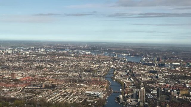 High-altitude city view over Amsterdam showing dense urban neighborhoods, waterways, and harbor areas under a pale blue sky.