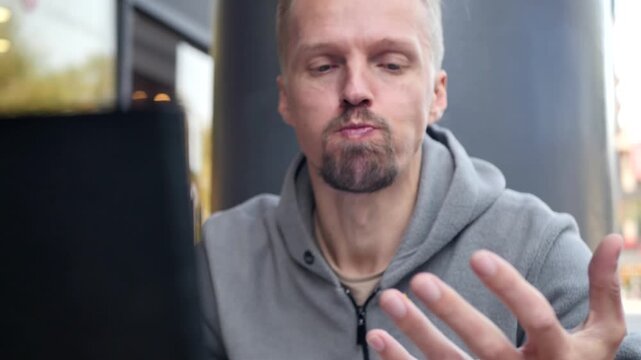 Young stylish blond man works on laptop, eats hamburger and speaks on online video call via laptop camera. Remote work of programmer and IT specialist without lunch break.
