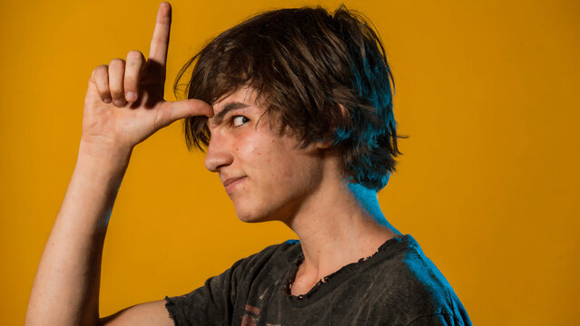 Profile of a teenage boy making a "loser" hand gesture on his forehead against a bright yellow background