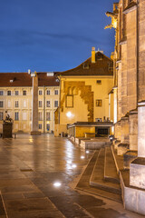 Fototapeta premium The Mrakotin Monolith stands tall at Prague Castle, surrounded by historical buildings, as warm light casts dramatic shadows on the cobblestone ground under the night sky.