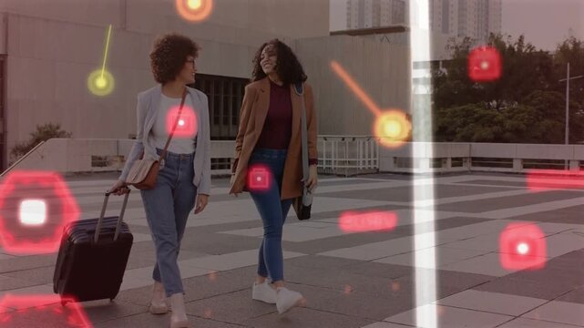 Walking two women in coats smiling and talking across plaza, pulling suitcase with red graphics