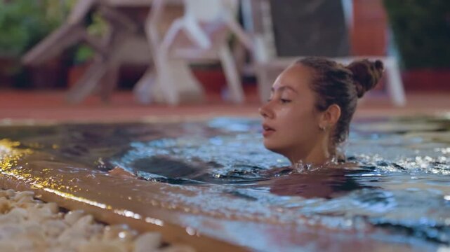 Evening pool relax at island resort, woman pausing at pool edge to catch breath, wet hair bun, ambient lights and potted foliage, shallow ripples and reflective water surface conveying calm rest