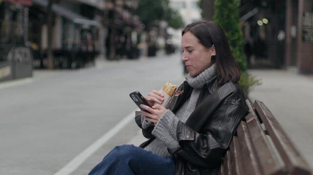 Woman eating a sandwich and using a smartphone on a city bench