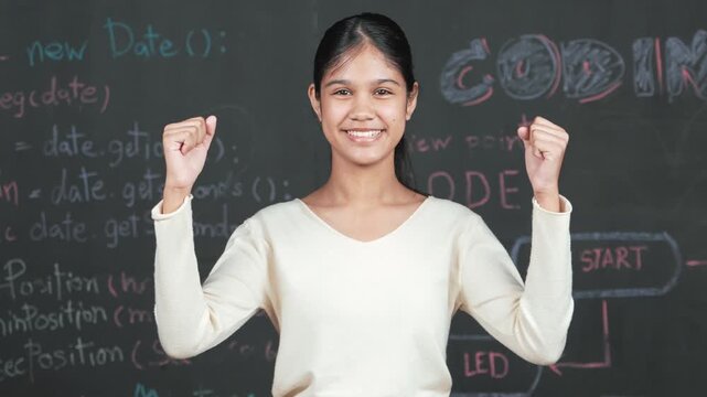 Young student celebrate her successful plan while raise her arm. Happy teenager looking at camera while standing at blackboard with engineering code and prompt written in STEM class. Edification