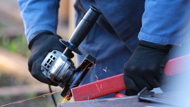 The worker will make an angle grinder for processing a metal part, bright sparks fly to the sides. The concept of industrial labor, repair, construction of safety equipment.