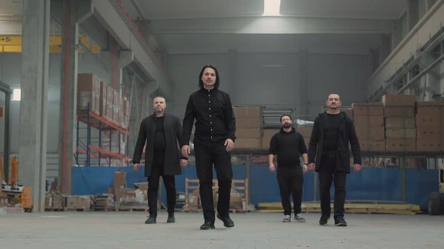 Group of four men in black clothes walking forward through a large warehouse a male rock band moving together on the concrete floor past storage racks.