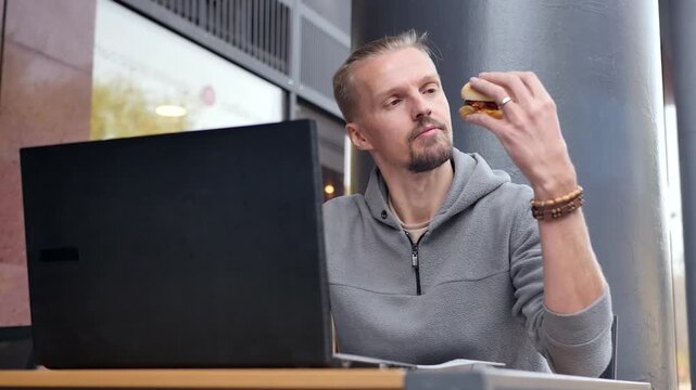 Young slim man attentively studying and evaluating looks at hamburger holding it in hand and preparing for lunch. Person works with laptop and eats. Lunch break in outdoor cafe. Fast food at work.