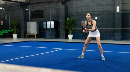 Obraz premium Female athlete holding padel racket ready on cobalt-blue court wearing gray tank top, copy space
