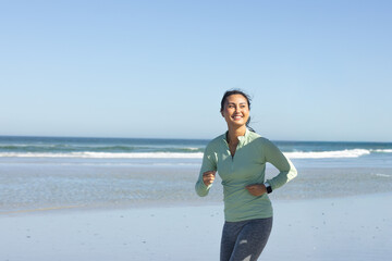 Obraz premium Mid-adult Asian woman jogging along wet sand by sea in green top checking smartwatch, copy space