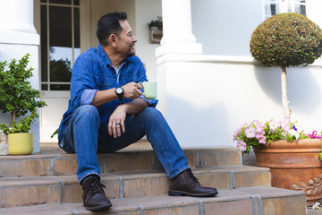 Middle-aged Asian man sitting on porch steps in denim shirt holding light green mug, looking left © wavebreak3