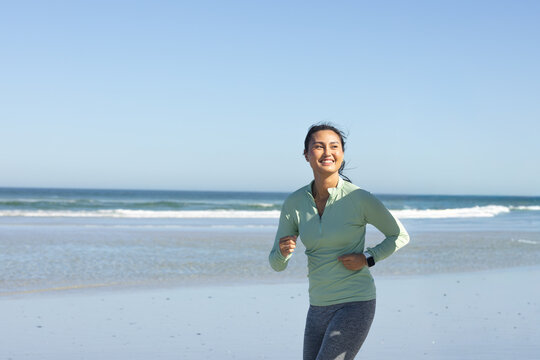 Mid-adult Asian woman jogging along wet sand by sea in green top checking smartwatch, copy space