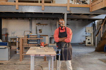 Obraz premium African American male carpenter posing arms crossed at shop bench goggles sander clamps, copy space