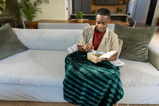 African American man sitting on light gray sofa, eating takeout with chopsticks under green blanket