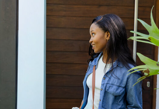 African American woman standing by wood-paneled door, wearing denim, holding bottle, copy space