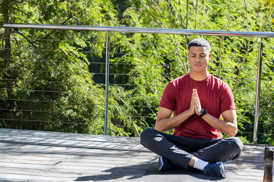 Man meditating on yoga mat on wooden deck wearing red tee, smartwatch, travel bottle, copy space