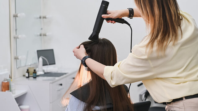 A hairdresser blow-dries a client's long, natural, dark hair with a round brush and a styler at a salon. Professional hair styling and care at the hair salon.