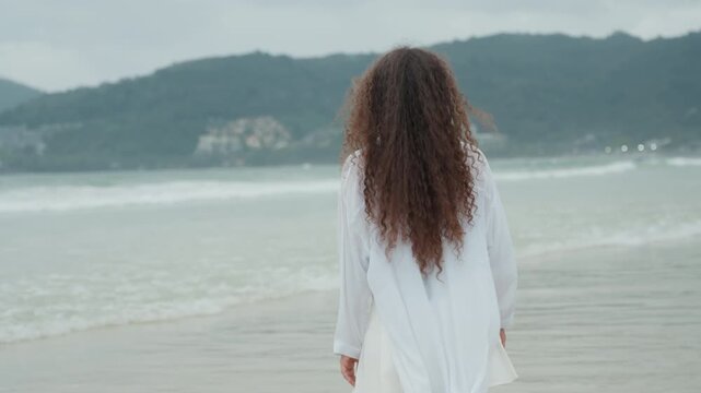 Biracial woman facing island ocean horizon, flowing white coverup, long curly hair, gentle surf and distant islets, empty sand, reflective walking, slow backtracking shot, melancholic calm