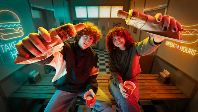 Two friends holding hot dogs toward camera in neon lit retro diner interior. Concept of social dining promotion, group offer advertising, fast food branding and shared meal marketing.