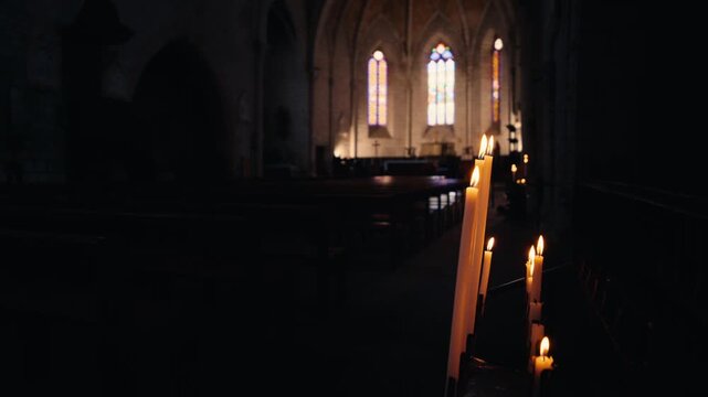 Lit candles glow in the dark interior of the church in Monpazier, Dordogne. Stained glass windows illuminate the background. Slow motion traveling shot.