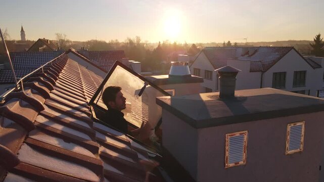 Man drinks tea while leaning out of an attic window on a frosty winter morning at sunrise. Cozy everyday moment above a quiet residential neighborhood with snowy rooftops and warm sunlight.