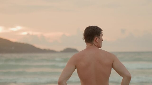 Sunset island coastline with white man hands on hips, strong stance facing horizon, muscular silhouette against warm sky and rolling surf, moment of resolve and preparedness as evening tide laps