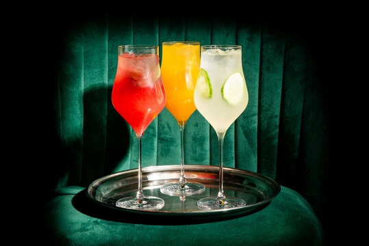 Three colorful cocktails (red, orange, and white) in tall glasses on a silver tray against a dark green velvet background.