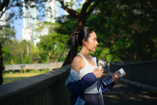 Woman holding portable fan and water bottle after workout, cooling down