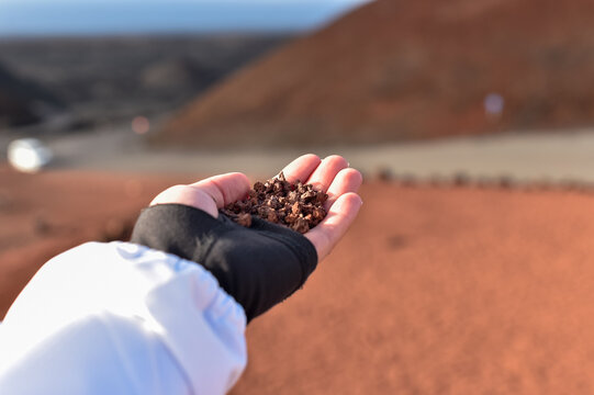 A person holds a handful of red volcanic lapilli rocks in an open palm. The background shows a blurred red volcanic landscape and hills in Lanzarote.