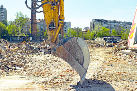 Excavator bucket resting on rubble at active demolition site with urban background. Illustrates heavy machinery operation, construction workflow, and controlled material removal during site clearance.