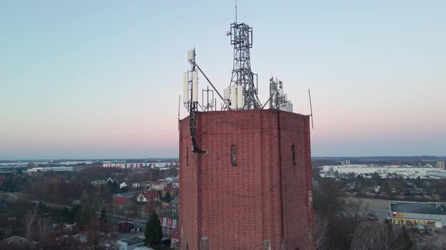Aerial view of a tall brick water tower with communication antennas above a small town under a soft dawn sky.